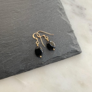 May include: A pair of dangle earrings featuring smooth, oval-shaped black stones suspended from gold-toned metal ear wires. Each earring consists of a single polished black bead attached to a delicate gold wire hook with a small gold bead accent.