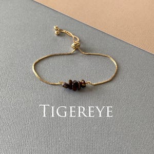 May include: A gold-toned adjustable bracelet featuring a delicate box chain and a central cluster of small, polished brown tiger eye gemstone chips. The word TIGEREYE is printed in white capital letters below the jewelry piece.