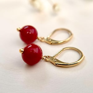 May include: A pair of gold-tone leverback earrings featuring round, polished red beads. Each earring consists of a single spherical red bead suspended from a metallic gold-tone leverback mechanism, showcasing a simple and classic design.