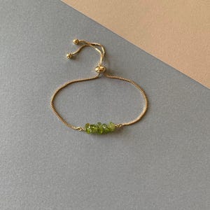 Puede incluir: Una pulsera ajustable en tono dorado con una cadena fina de caja y una fila central de cuatro pequeñas piedras de peridoto verde, crudas y translúcidas. La pulsera incluye un mecanismo de cuenta deslizante con dos extremos colgantes.
