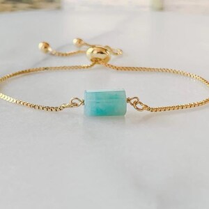 May include: A gold-toned adjustable bracelet featuring a single rectangular, light blue-green stone bead at the center. The bracelet is constructed with a delicate box chain and includes a sliding bead mechanism to adjust the fit of the jewelry.