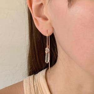 Puede incluir: Un pendiente de hilo de oro rosa con una delicada cadena de eslabones cuadrados que atraviesa el lóbulo. Un cristal transparente de corte bruto en forma de punta cuelga de la cadena, sujeto por una pequeña tapa de metal de oro rosa.