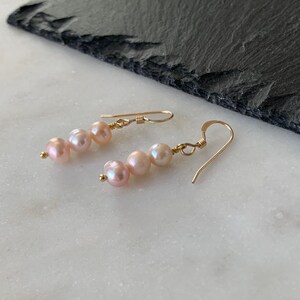 May include: A pair of drop earrings featuring three small, round, light pink pearls arranged vertically on a thin gold wire. The earrings are attached to gold-toned ear hooks, creating a delicate and simple design that hangs downward from the wire.