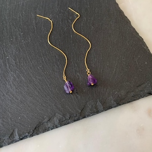 Puede incluir: Un par de pendientes de hilo dorados con cadenas largas y onduladas que terminan en pequeñas piedras preciosas de amatista de color púrpura intenso e irregular. Los pendientes presentan un diseño de cadena metálica fina y delicada.
