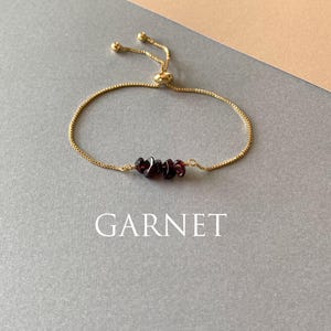 May include: A gold-toned adjustable bracelet featuring a delicate chain and three small, dark red garnet gemstone chips at the center. The bracelet includes a sliding bead mechanism for sizing and the word GARNET is printed below the jewelry.