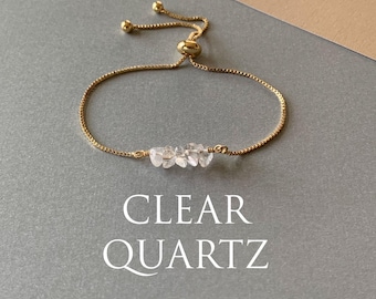May include: A gold-toned adjustable slider bracelet featuring a delicate box chain and a central row of small, raw clear quartz crystal chips. The text CLEAR QUARTZ is printed in white below the bracelet, which includes two small gold beads.