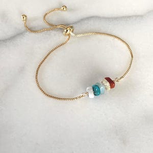 May include: A gold-tone adjustable slider bracelet featuring a delicate chain and a central cluster of four small, polished gemstone beads in white, turquoise, pale yellow, and deep red, finished with two small gold-tone beads at the ends.