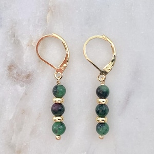 May include: A pair of gold leverback earrings featuring a vertical drop design. Each earring consists of three round, mottled green and dark red gemstone beads separated by two small, smooth, polished gold-toned metal spacer beads.