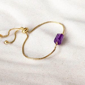 May include: A gold-toned adjustable bracelet featuring a thin box chain and a single cylindrical purple gemstone bead. The bracelet includes a sliding bead mechanism for size adjustment, finished with two small gold-toned spheres at the ends.