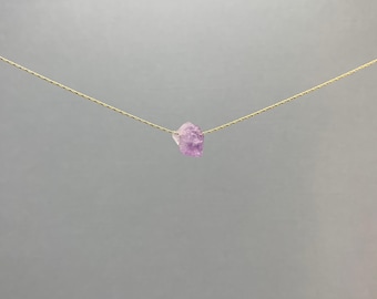 May include: A delicate gold-tone chain necklace featuring a single, small, raw, light purple amethyst crystal pendant suspended in the center. The chain is thin and metallic, providing a simple and clean look with the natural stone as the focal point.