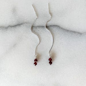 May include: A pair of silver threader earrings featuring a delicate box chain design. Each earring terminates in two small, faceted deep red garnet beads, creating a subtle and elegant drop effect that hangs vertically from the silver chain.