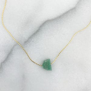 May include: A delicate gold chain necklace featuring a single, small, raw, light green aventurine stone pendant. The stone has an irregular, angular shape and is suspended centrally on the thin, metallic gold-coloured link chain.