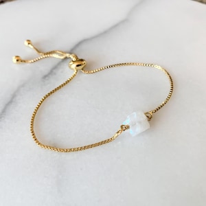 Op de afbeelding: Een goudkleurige armband met een fijne box-ketting en een enkele, doorschijnende, iriserende, vierkant geslepen steen in het midden. De armband is voorzien van een verstelbaar schuifmechanisme met twee kleine goudkleurige bolletjes aan de uiteinden.
