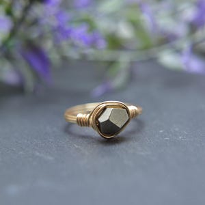 May include: A gold-toned wire-wrapped ring featuring a single, small, metallic gray, multifaceted pyrite stone at the center. The band is constructed from thin, coiled gold-colored wire that secures the stone in a simple, open-frame setting.