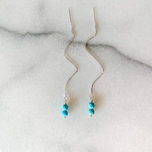 Puede incluir: Un par de pendientes de hilo de plata con un diseño de cadena fina. Cada pendiente termina en dos pequeñas cuentas facetadas de color turquesa, creando un estilo colgante sencillo y elegante que cuelga verticalmente del hilo.