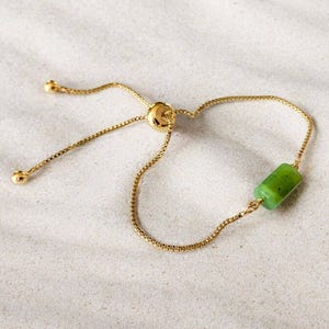 Op de afbeelding: Een verstelbare goudkleurige armband met een dunne boxketting en een enkele cilindrische groene stenen kraal. De armband bevat een schuifkraalmechanisme voor maatverstelling, afgewerkt met twee kleine goudkleurige bolletjes aan de uiteinden.