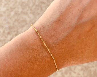 May include: A delicate gold chain bracelet featuring small, evenly spaced gold beads along the length of the chain. The bracelet is thin and minimalist in design, showcasing a simple link structure with polished spherical accents throughout the piece.