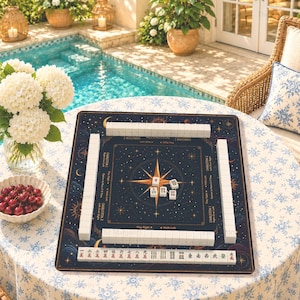 May include: A square mahjong mat featuring a dark blue celestial design with gold stars, moons, and a central compass rose. White tiles are arranged in rows along the edges and in the center. The mat includes printed text for game instructions.