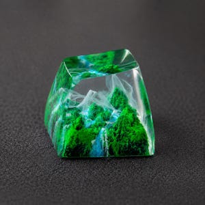May include: A translucent resin mechanical keyboard keycap featuring a miniature landscape design. The interior displays vibrant green mossy mountain peaks, deep blue water elements, and wispy white cloud-like formations encased in clear material.