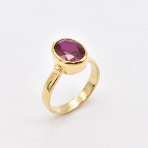 Puede incluir: Un anillo de oro con una gran gema rosa oscuro de forma ovalada. La banda y el engaste del anillo son de oro pulido. La gema es el punto focal, reflejando un color rico. El anillo está sobre un fondo blanco liso. Es una pieza de joyería.