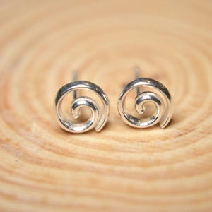 Pode incluir: Um par de brincos prateados com um design em espiral. Os brincos são exibidos em uma superfície de madeira marrom clara. Cada brinco tem uma forma giratória.