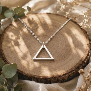 Puede incluir: Un collar de tono plateado que presenta un colgante de triángulo abierto y sencillo suspendido de una cadena delicada. El colgante es pulido y liso, refleja la luz, y la cadena consta de pequeños eslabones circulares uniformes unidos en la punta.