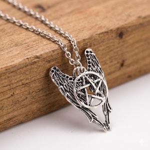 Op de afbeelding: Zilverkleurige hangerketting met een pentagram in een cirkel, geflankeerd door gestileerde vleugels. De ketting is delicaat. De ketting wordt gepresenteerd op een houten oppervlak.