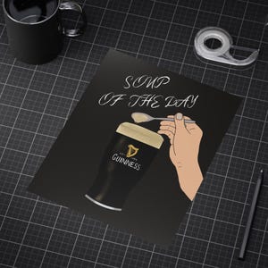 May include: Black poster with the text "Soup of the Day" above a pint of Guinness beer. A hand is depicted holding a spoon with a dollop of foam, above the beer. A black mug, tape dispenser, and pencil are also visible.