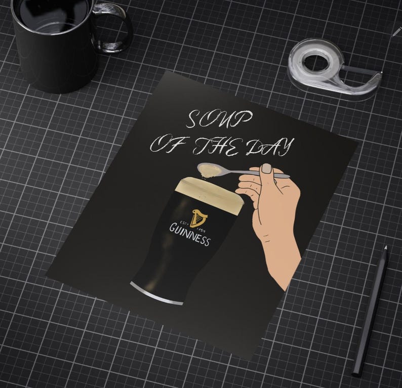 May include: Black poster with the text "Soup of the Day" above a pint of Guinness beer. A hand is depicted holding a spoon with a dollop of foam, above the beer. A black mug, tape dispenser, and pencil are also visible.