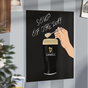 May include: A black poster with the words "SOUP OF THE DAY" in white script. A hand holds a spoon over a pint of Guinness. The Guinness glass has the brand logo on it.