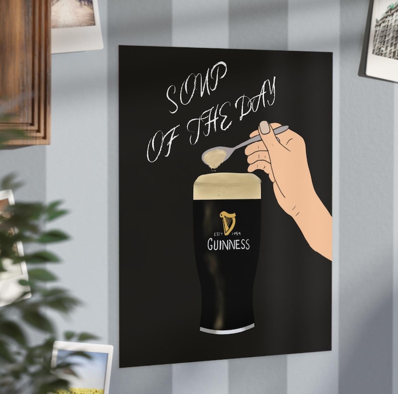May include: A black poster with the words "SOUP OF THE DAY" in white script. A hand holds a spoon over a pint of Guinness. The Guinness glass has the brand logo on it.