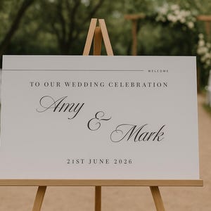 Peut inclure: Un panneau de mariage blanc sur un chevalet en bois. Le panneau indique « TO OUR WEDDING CELEBRATION » avec les noms « Amy & Mark » et la date « 21 juin 2026 ». L'arrière-plan montre un chemin et des chaises blanches.