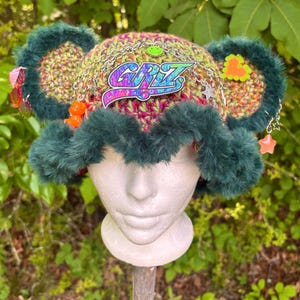 May include: A colorful crocheted hat with green faux fur trim and ear-shaped accents. The hat features the word "CRZ" in a vibrant, multi-colored design, along with decorative chains, charms, and beads. The hat is displayed on a white mannequin head.