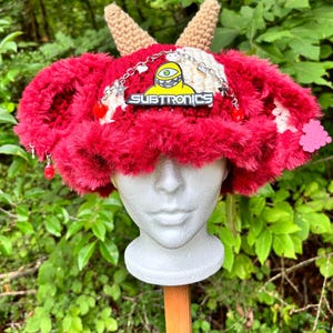 May include: A red and white fuzzy crochet hat featuring two tan crocheted horns on top. The hat is decorated with a silver chain, small charms, and a yellow cyclops character patch with the text SUBTRONICS in black and white lettering.