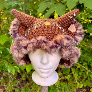May include: A brown crocheted hat with two upward-curving horns, adorned with a faux fur trim in shades of brown and purple. The hat features a decorative chain with small charms and a flower-shaped embellishment. The hat is displayed on a white mannequin head.