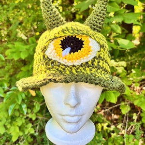 May include: A crochet bucket hat in variegated shades of lime and olive green, featuring two pointed ears on top and a large, centered eye motif. The eye has a white sclera, a bright yellow iris, a black pupil, and a small light blue accent.