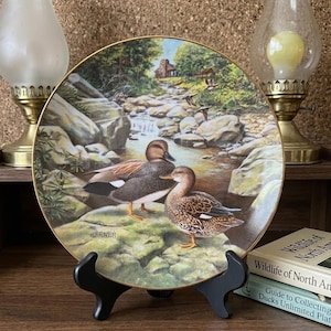 May include: A decorative plate featuring a detailed painting of two ducks near a stream with a small cabin in the background. The plate has a gold rim and is displayed on a black stand. The artist's name, JERNER, is visible.