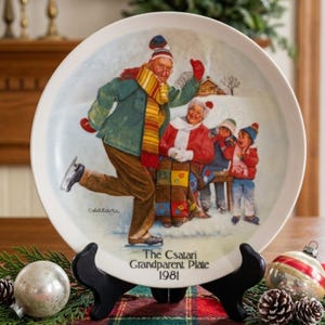 May include: A decorative plate with a winter scene of a family ice skating. The plate has a white border and the text "The Csatari Grandparent Plate 1981". The illustration shows a man skating, a woman seated, and two children. The plate is on a black stand.