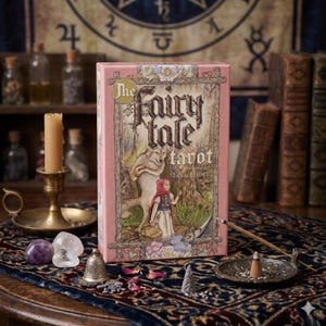 Puede incluir: Una caja de tarot rosa titulada "The Fairy Tale Tarot" con una ilustración de Caperucita Roja y un lobo. La escena incluye cristales, una vela, incienso y libros antiguos, creando una estética mística.