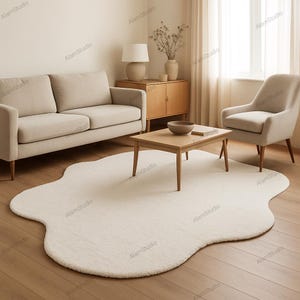 Op de afbeelding: Een crèmekleurig, wolk-vormig vloerkleed ligt op een houten vloer in een woonkamer. Het vloerkleed wordt omringd door een beige bank, een fauteuil, een houten salontafel en een houten kast. Een lamp en een vaas staan op de kast.