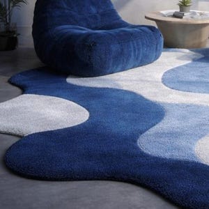 May include: A modern, abstract area rug in shades of blue and white. The rug features a wavy, organic design with varying tones of blue, creating a sense of depth and movement. The rug is placed on a gray concrete floor, with a blue beanbag chair in the background.