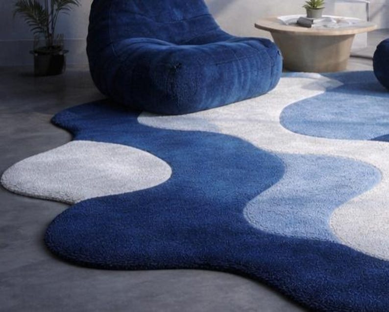 May include: A modern, abstract area rug in shades of blue and white. The rug features a wavy, organic design with varying tones of blue, creating a sense of depth and movement. The rug is placed on a gray concrete floor, with a blue beanbag chair in the background.