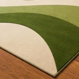 May include: A green, cream, and light green area rug with a wave design. The rug is placed on a hardwood floor. The rug's design features curved lines and a soft texture, adding a decorative element to the room.