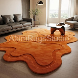 Könnte beinhalten: Ein orangefarbener, abstrakt geformter Teppich mit einem geschichteten Design. Der Teppich liegt auf einem Holzboden in einem Wohnzimmer. Der Teppich hat eine einzigartige, fließende Form und eine warme Farbe. Der Text "Alam Rugs Studio" ist auf dem Teppich zu sehen.