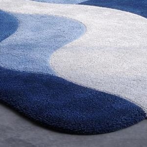 May include: A close-up of a blue and white area rug with a wavy design. The rug features shades of dark blue, light blue, and white, creating a textured, flowing pattern. The rug is made of a soft, plush material.