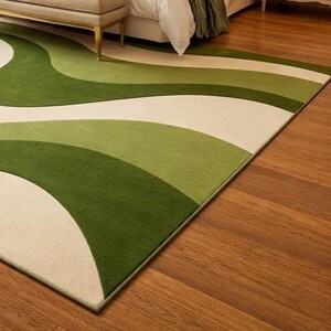 May include: A green and cream-colored area rug with a wavy design. The rug features shades of green and cream in a flowing pattern. The rug is rectangular and appears to be made of a soft material. The rug is placed on a hardwood floor.