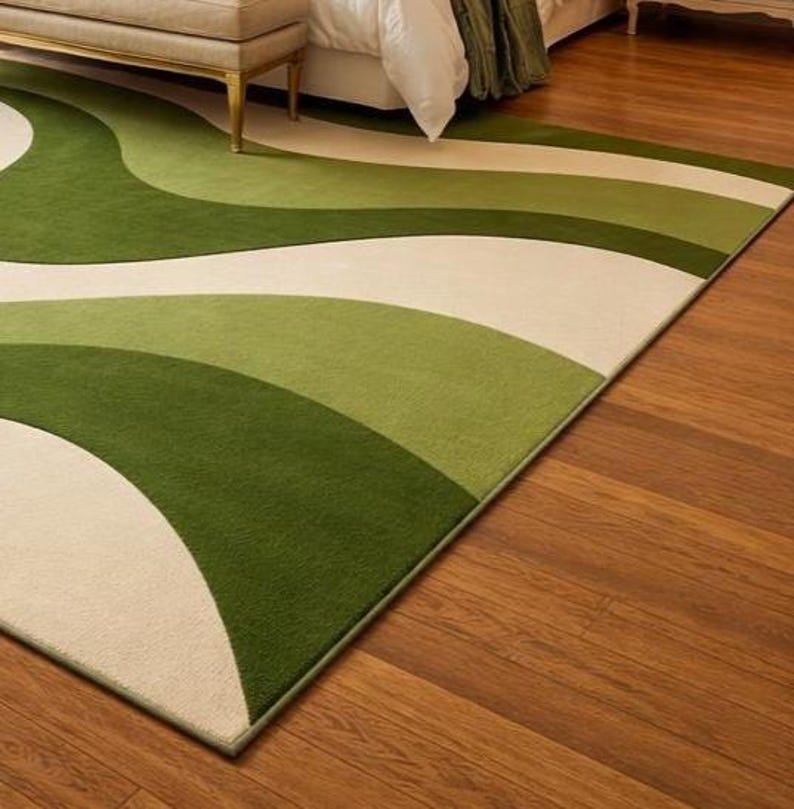 May include: A green and cream-colored area rug with a wavy design. The rug features shades of green and cream in a flowing pattern. The rug is rectangular and appears to be made of a soft material. The rug is placed on a hardwood floor.