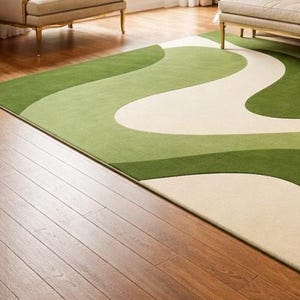 May include: A rectangular area rug with a flowing, abstract design in shades of green and cream. The rug is placed on a hardwood floor, with a glimpse of furniture in the background. The rug's pattern creates a sense of movement.