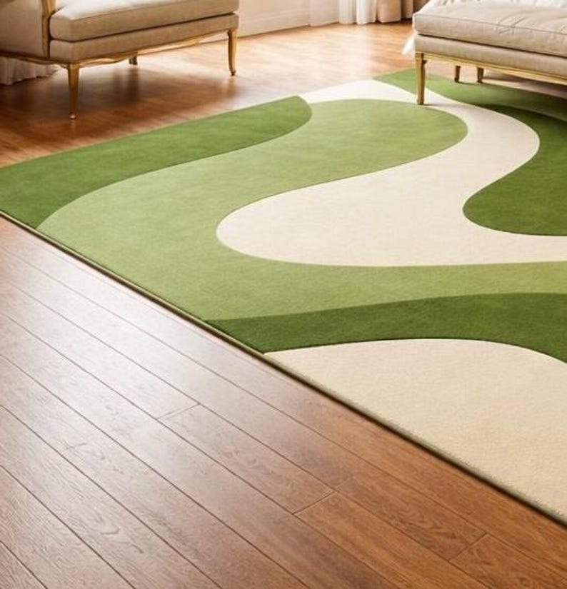 May include: A rectangular area rug with a flowing, abstract design in shades of green and cream. The rug is placed on a hardwood floor, with a glimpse of furniture in the background. The rug's pattern creates a sense of movement.