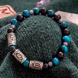 May include: A beaded bracelet featuring alternating round blue and black stones, interspersed with silver-toned cylindrical metal beads engraved with runic symbols. The bracelet is arranged in a circular shape, showcasing a polished finish.